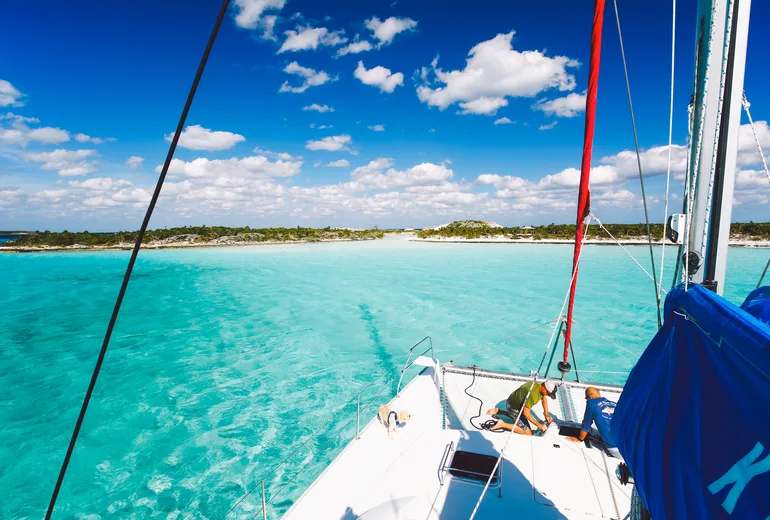 A sailboat foredeck with two crew approaching a low sandy cay over turquoise banks
