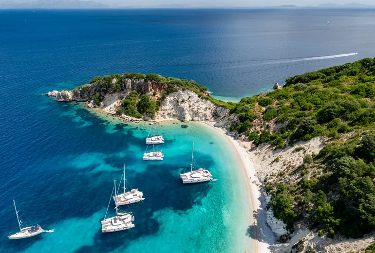 Aerial of charter yachts anchored off a white pebble cove on a green Ionian islet