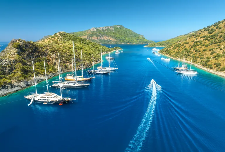 Aerial of gulets and yachts anchored in a narrow turquoise channel between pine hills, Türkiye