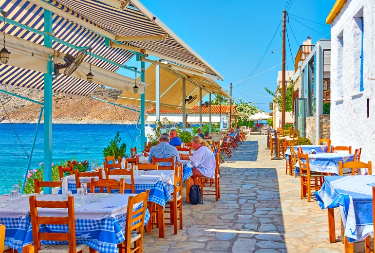 Blue-checked taverna tables along a stone waterfront promenade with a few diners, Greece