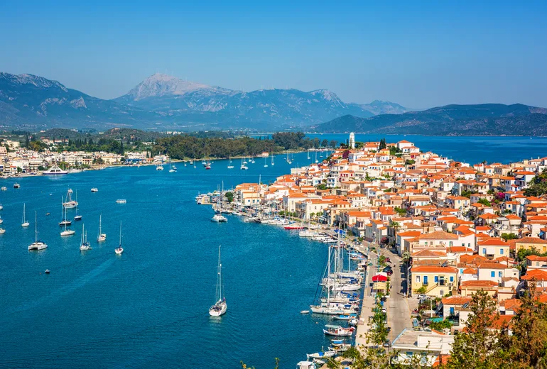 Poros town waterfront with anchored sailboats and a mainland mountain backdrop, Greece