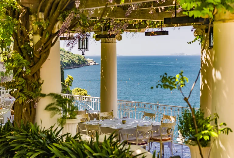 Restaurant terrace under a wisteria-covered pergola with columns overlooking the sea, Italy