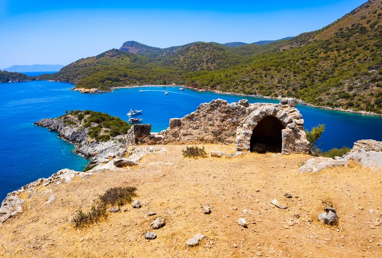 Ruined stone chapel arch on an island hilltop above a turquoise anchorage and pine coast, Türkiye