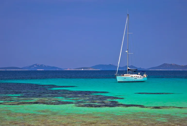 Sailboat anchored in turquoise shallows with low islands behind, Croatia