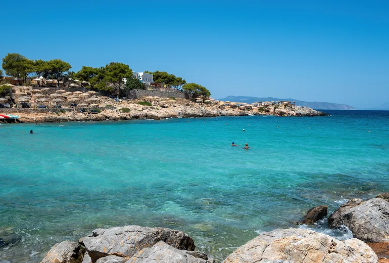 Swimmers in a turquoise rocky cove fringed by pines and sunbeds, Greece