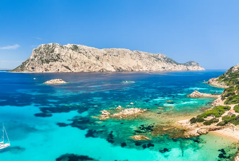 Tavolara island limestone massif over turquoise shallows with anchored sailboat, Sardinia