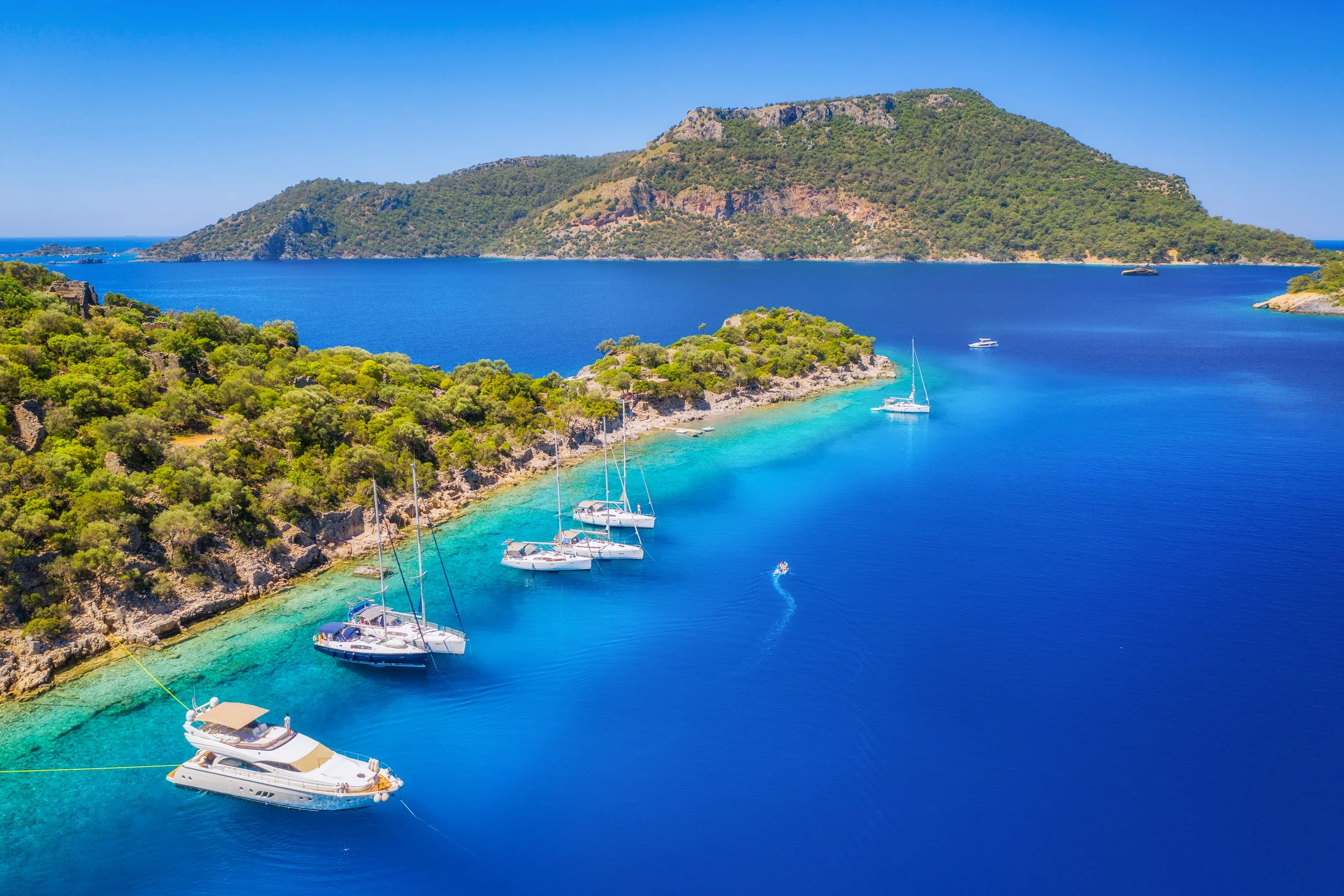 Aerial of yachts moored along pine shore in turquoise bay facing island, Turkey