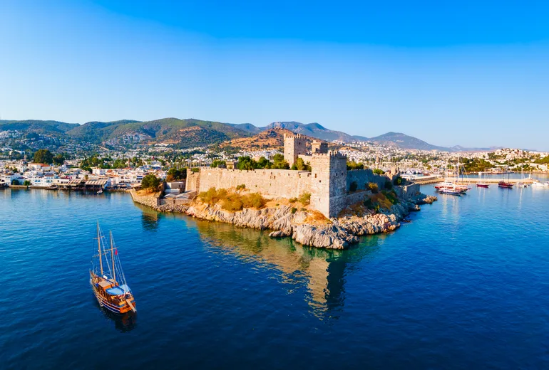 Wooden gulet sailing past Bodrum castle walls with town and hills behind